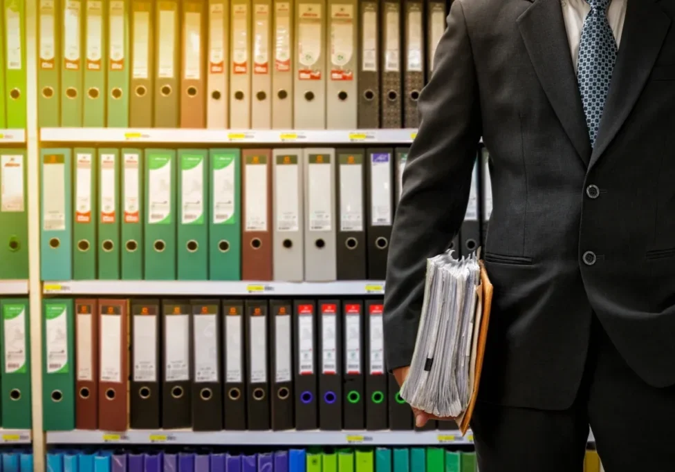 A man holding a briefcase stands beside shelves filled with colorful binders.